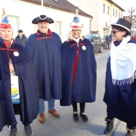 Vier Personen in traditionellen Kostümen stehen auf der Straße. Zwei tragen blaue Roben mit roten Säumen, eine hat einen dreifarbigen Hut und die andere einen schwarzen Hut. Die verbleibenden beiden tragen weiße Umhänge. Ein Auto ist hinter ihnen geparkt.