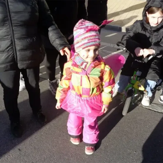 Ein kleines Mädchen in einem bunten Outfit steht auf der Straße und lächelt. Dahinter stehen zwei Erwachsene und ein Kind auf einem Fahrrad. Das Kind trägt einen Helm.