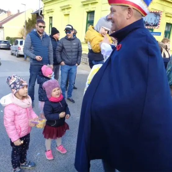 Menschen versammeln sich auf einer Straße mit einem Mann in Kostüm. Zwei junge Mädchen stehen neben ihm, eine hält einen Korb. Dahinter beobachten eine Gruppe Erwachsener, einige mit Mützen. Im Hintergrund steht ein gelbes Gebäude und geparkte Autos.