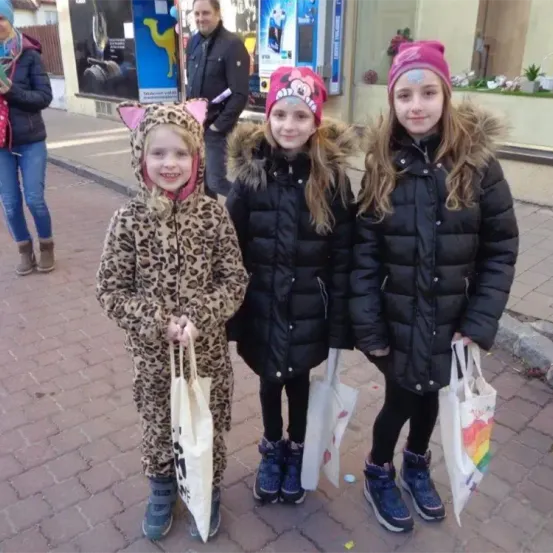 Drei junge Mädchen stehen auf einer gepflasterten Straße. Das Mädchen auf der linken Seite trägt ein Leoparden-Kostüm und die anderen beiden schwarze Wintermäntel. Sie halten alle Einkaufstüten. Dahinter ist ein verschwommenes Bild eines Mannes und einer Frau, die vorbeigehen.