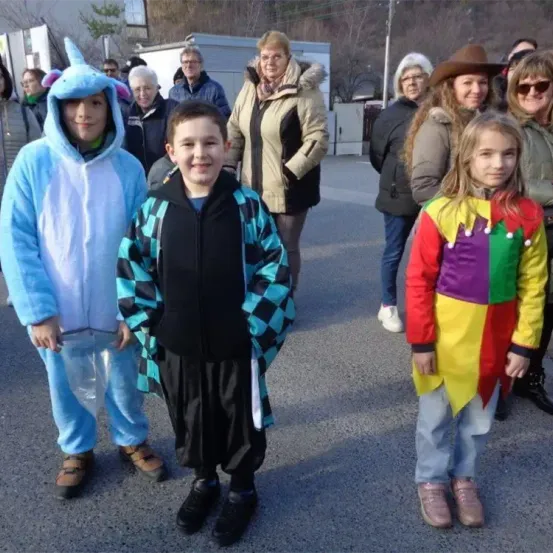 Eine Gruppe von Kindern und Erwachsenen in Kostümen steht auf einem Außengelände. Die Kinder sind in Tier- und bunte Kostüme gekleidet. Hinter ihnen stehen Erwachsene und beobachten. Sie befinden sich auf einem Parkplatz.