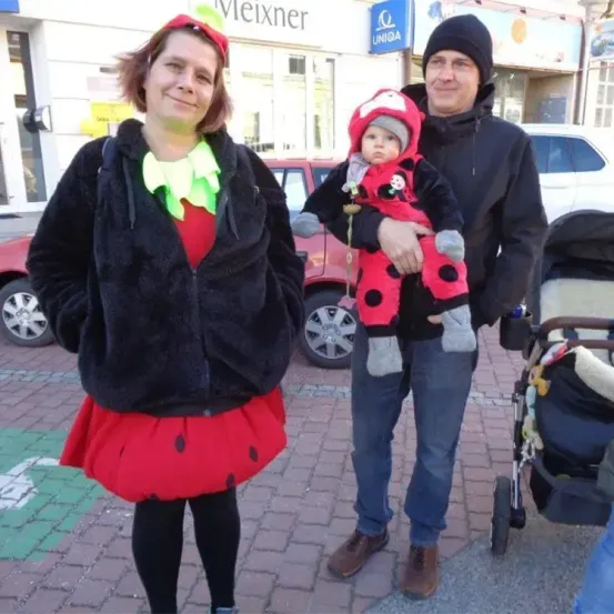 Eine dreiköpfige Familie steht auf einer gepflasterten Straße, alle in Kostümen. Eine Frau in einem roten Marienkäferkleid, ein Mann in einem schwarzen Jackett und ein Baby in einem Marienkäfer-Kostüm. Hinter ihnen steht ein roter Wagen und ein Kinderwagen.