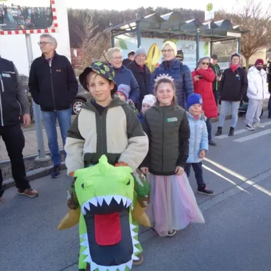 Eine Gruppe von Menschen in Festkostümen, darunter ein Junge in einem Dinosaurierkostüm und ein Mädchen in einem rosa Tutu, gehen auf der Straße.