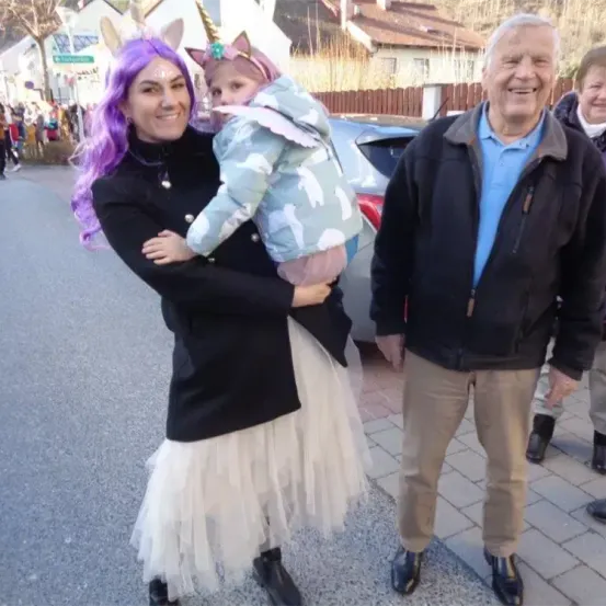 Eine Frau in einem Einhornkostüm hält ein Kind in einem Kostüm auf der Straße. Ein älterer Mann neben ihr lächelt.
