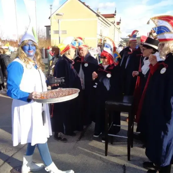 Eine Frau in einem Kostüm mit blauer Gesichtsbemalung hält einen Tablett mit Essen. Sie ist von Leuten in bunten Hüten umgeben.