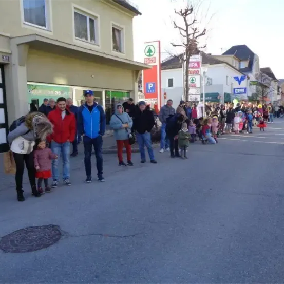 Eine Gruppe von Menschen wartet am Straßenrand, mit mehreren Kindern in Kostümen, die in einer Reihe stehen. Im Hintergrund befinden sich Gebäude mit Schildern und ein Baum.