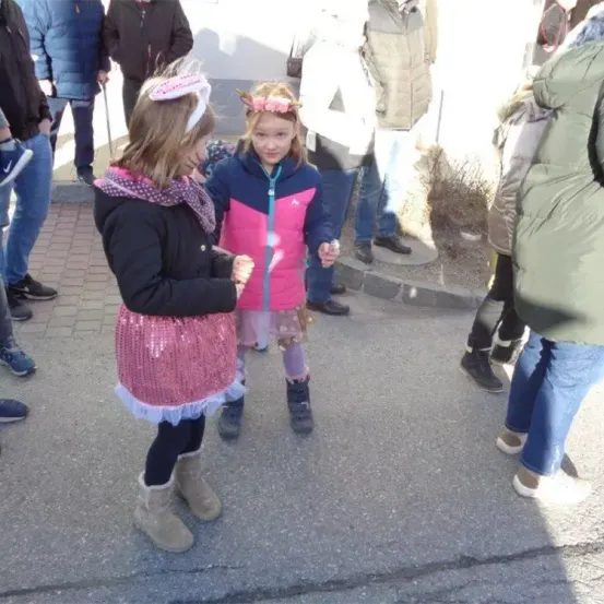 Zwei junge Mädchen in Winterkleidung stehen auf der Straße, eines in einem rosa Rock und das andere in einer rosa und blauen Jacke, beide halten sich an den Händen.