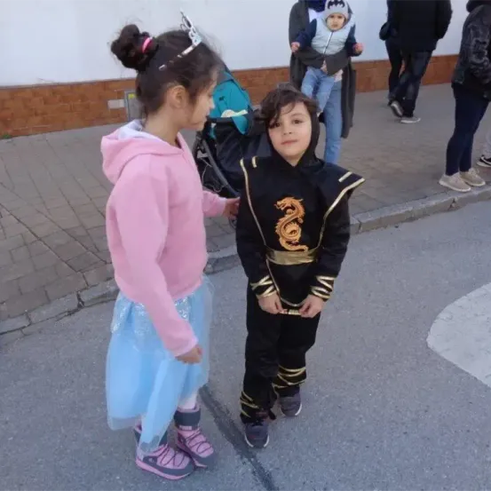 Zwei Kinder, ein Mädchen in Pink und ein Junge in einem Drachenkostüm, stehen auf einer Straße. Hinter ihnen ist eine Person mit einem Baby im Kinderwagen und zwei gehende Personen zu sehen.