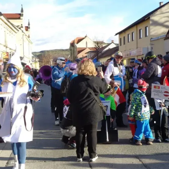 Eine Straßenszene mit einer Menschenmenge in Kostümen und Skiausrüstung, einige halten Instrumente, und eine Person macht ein Foto mit einer Kamera. Gebäude säumen die Straße.