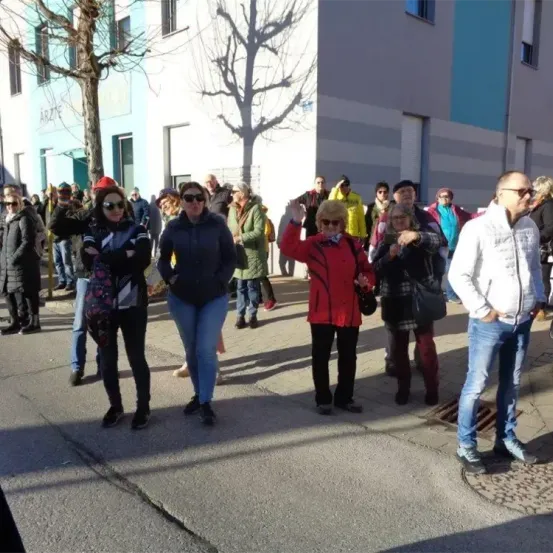Eine Gruppe von Menschen in warmer Kleidung steht auf einer Straße. Einige winken, andere unterhalten sich. Im Hintergrund befindet sich ein Gebäude mit einem Baum und Schatten.
