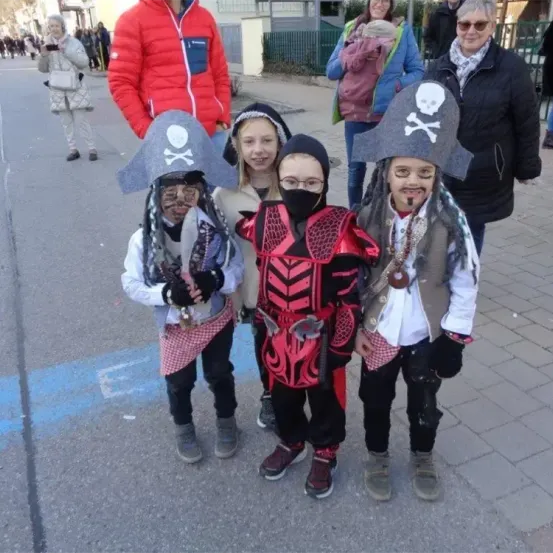 Vier Kinder, verkleidet als Piraten, stehen auf einer Straße, eines mit einem Schwert, ein anderes mit einer Maske und ein Mädchen in einem rot-schwarzen Kostüm. Dahinter stehen Erwachsene und beobachten, einer trägt ein Baby.