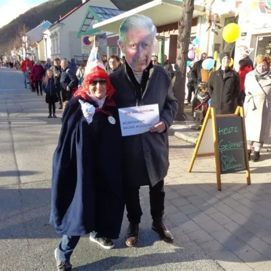 Eine Person, die als älterer Mann gekleidet ist, und eine andere in einer roten Perücke stehen auf einer Straße, halten ein Schild und lächeln für ein Foto.