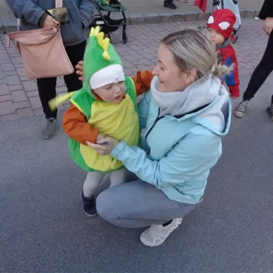 Eine Frau in einer hellblauen Jacke kniet sich hin und hält ein Kind in einem Dinosaurierkostüm. Hinter ihnen trägt ein Mann eine rosa Handtasche, und ein Kind in einem Spider-Man-Kostüm steht in der Nähe.