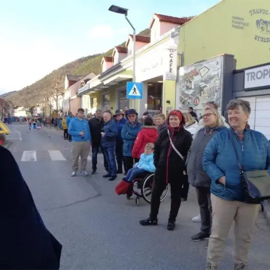 Eine Gruppe von Menschen versammelt sich auf einer Straße, mit einem Gebäude, das als Tropic und ein anderes als Cafe beschriftet ist. Eine Person im Rollstuhl ist von einigen anderen umgeben.