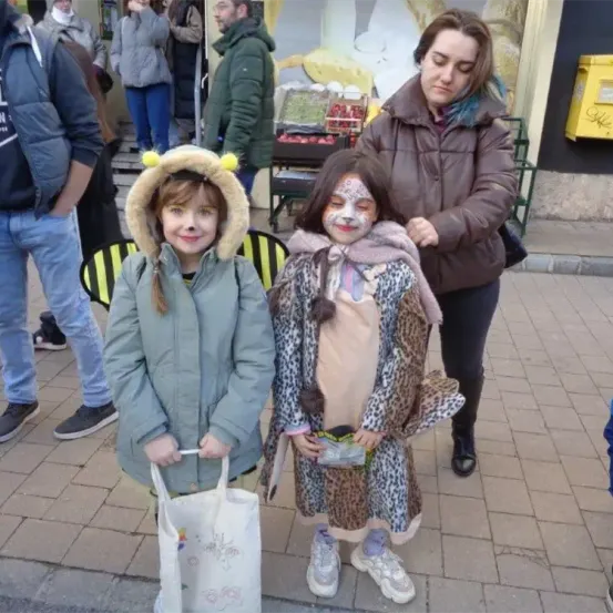Zwei Mädchen in Tierkostümen stehen auf einer gepflasterten Straße. Eines hält eine weiße Tasche. Hinter ihnen hält eine Frau in einer braunen Jacke und schwarzen Hosen den Arm eines Mädchens in Leopardenmuster. Leute stehen hinter ihnen, und eine gelbe Box befindet sich auf dem Bürgersteig.