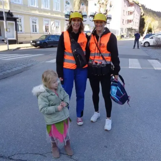 Zwei Frauen in orangefarbenen Westen stehen auf einer Stadtstraße mit einem kleinen Mädchen. Sie alle lächeln. Hinter ihnen sind geparkte Autos und eine Person, die geht.