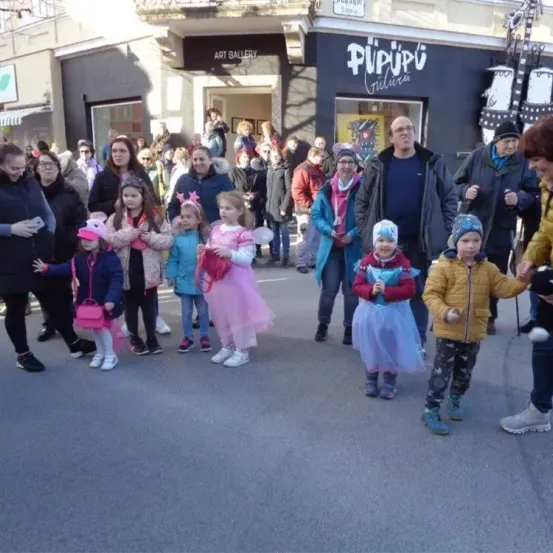 Eine Gruppe von Menschen, darunter Kinder in Kostümen, steht auf einer Straße, möglicherweise für eine Veranstaltung, mit Geschäften im Hintergrund.