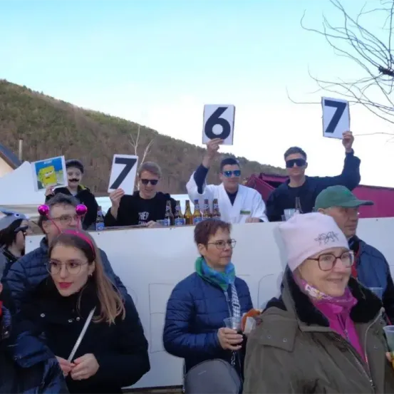 Eine Gruppe von Menschen in Kostümen und mit Brillen hält Schilder mit den Zahlen 6 und 7 hoch. Sie stehen vor einer weißen Wand mit Flaschen und Getränken obenauf.