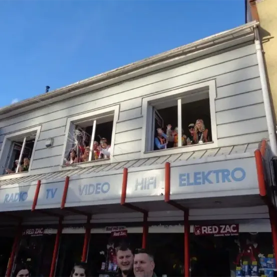 Ein Schaufenster zeigt 'RADIO', 'TV', 'VIDEO', 'HIFI' und 'ELEKTRO' Schilder. Leute im oberen Fenster winken und es stehen zwei Männer vor dem Laden.
