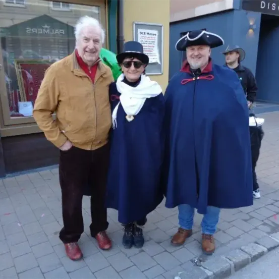 Drei Erwachsene stehen lächelnd auf einer Straße. Einer trägt eine braune Jacke, ein anderer einen blauen Umhang und der dritte einen schwarzen Hut. Hinter ihnen befindet sich ein Geschäft mit einem Schild.