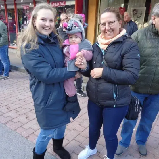 Zwei Frauen und ein Baby in einem Koala-Kostüm stehen auf einem gepflasterten Gehweg. Hinter ihnen stehen ein paar Leute, und im Hintergrund sind Geschäfte mit einigen ausgestellten Produkten.