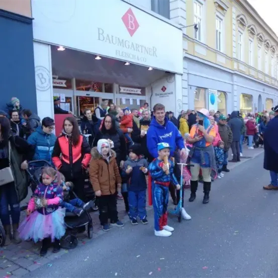 Eine Gruppe von Menschen steht auf der Straße vor einem Baumgartner Fleisch & Co-Geschäft, einige in Kostümen und mit Eis am Stiel.
