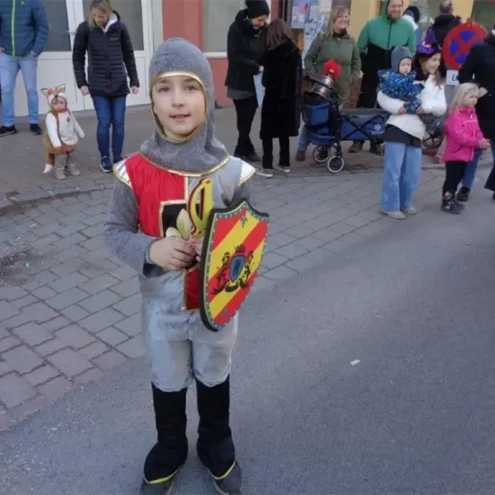 Ein Kind, das als Ritter verkleidet ist, hält einen Schild und ein Schwert in der Hand. Hinter ihnen gehen und stehen Leute, darunter eine Frau mit einem Baby im Kinderwagen.