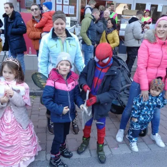 Eine Gruppe von Kindern und Erwachsenen, einige in Kostümen, stehen auf einem Bürgersteig mit Ziegelpflaster. Ein Kind, verkleidet als Spider-Man, hält einen Beutel, während ein anderes in einem rosa Kleid eine Krone trägt. Im Hintergrund sind Menschen in Winterkleidung zu sehen.