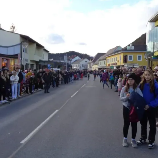 Eine Menschenmenge steht entlang der Straße, wahrscheinlich für eine Parade. Einige tragen Hüte und Brillen, während andere Taschen tragen. Eine Frau mit Brille und Rucksack steht in der Mitte. Gebäude säumen die Straße.