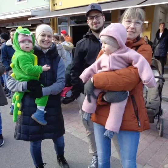 Eine vierköpfige Familie, darunter zwei Kinder in Kostümen, steht auf einer Stadtstraße mit einer Menschenmenge im Hintergrund.