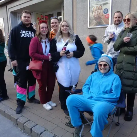 Eine Gruppe von Menschen in verschiedenen Kostümen steht auf einem Bürgersteig, lächelt und posiert für ein Foto. Hinter ihnen befindet sich ein Gebäude mit einem Poster an der Wand.
