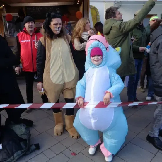 Eine Frau in einem Löwenkostüm und ein Kind in einem Einhornkostüm halten eine rot-weiße Barriere. Dahinter stehen mehrere Menschen, einige in Freizeitkleidung und andere in Winterkleidung.