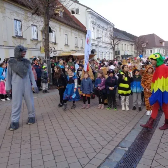 Eine große Menschenmenge, darunter Kinder in verschiedenen Kostümen, versammelt sich auf einem Stadtplatz. Einige tragen Schuhe, andere sind barfuß. Zwei Personen in Kostümen stehen rechts. Gebäude säumen die Straße dahinter.