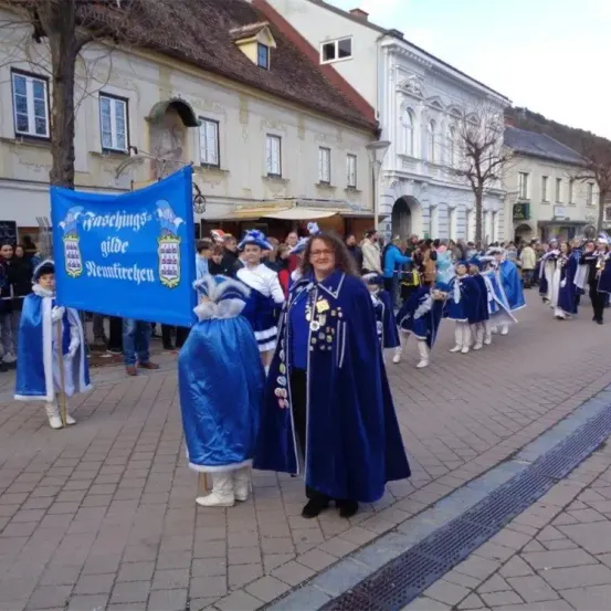 Eine Gruppe von Menschen in blauen Kostümen marschiert in einer Parade. Eine Frau in der Front hält ein blaues Banner mit weißem Text. Im Hintergrund befinden sich Bäume und Gebäude.