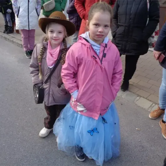 Zwei kleine Mädchen stehen auf der Straße, eines trägt eine rosa Jacke und einen blauen Rock mit Schmetterlingsmustern. Das andere Mädchen trägt einen braunen Hut, eine Schürze und eine braune Jacke. Hinter ihnen stehen mehrere Menschen.