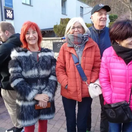 Vier Menschen stehen zusammen vor einem Gebäude mit einem Schild. Zwei Frauen tragen Pelzmäntel, eine in Schwarz und Weiß und die andere in Rot. Der Mann trägt eine blaue Jacke und eine Kappe. Die Frau rechts trägt eine pinke Jacke.