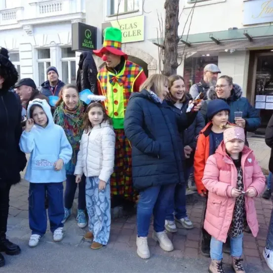 Eine Gruppe von Menschen, einschließlich Erwachsener und Kindern, steht auf einer gepflasterten Straße, wobei ein Mann als Clown verkleidet vor ihnen steht.