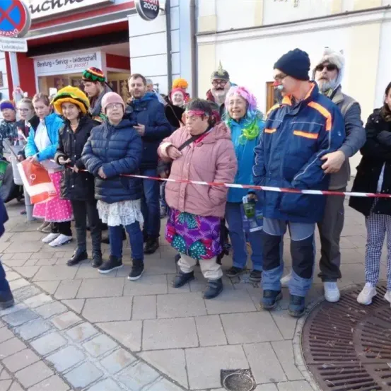 Eine Gruppe von Menschen in verschiedenen Kostümen steht auf einem gepflasterten Bürgersteig, einige halten Flaggen. Sie stehen vor einem Gebäude mit einem Schild und ein Mann in einer Mütze spricht zu ihnen.