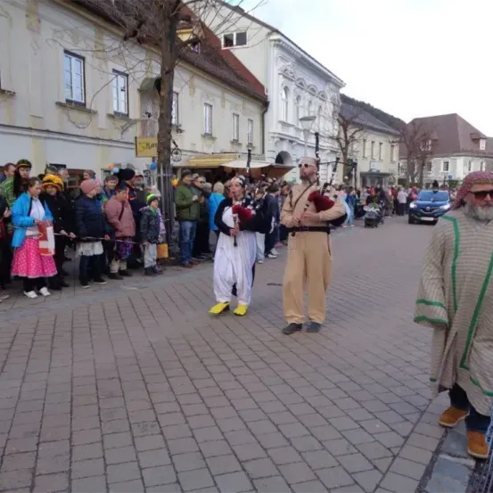 Ein Dudelsackspieler tritt auf einem Stadtplatz auf, während eine Menschenmenge zusieht. Gebäude und Bäume säumen die Straße.