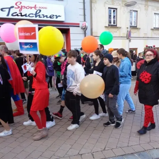 Eine Gruppe von Menschen in bunten Kostümen geht auf einer Straße vor einem Geschäft mit dem Schild Sport 2000. Sie halten Luftballons und Schilder in der Hand und nehmen wahrscheinlich an einer Parade teil.