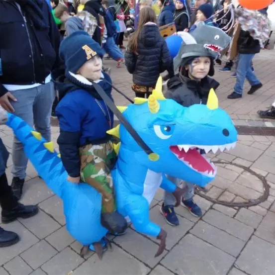 Zwei Kinder, verkleidet als blauer Dinosaurier und Hai, gehen auf einer gepflasterten Straße während einer Parade. Hinter ihnen gehen andere Leute, einige mit Luftballons.