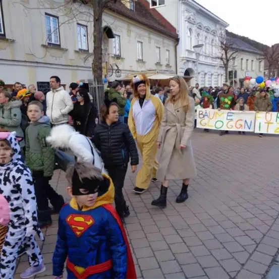Eine Gruppe von Menschen in Kostümen und Masken versammelt sich auf einem Stadtplatz. Im Hintergrund sind Gebäude, Bäume und Luftballons zu sehen. Ein Banner mit deutschem Text ist sichtbar.