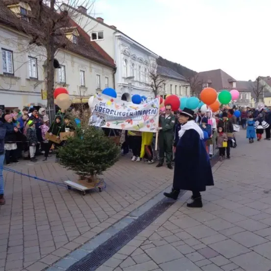 Eine Menschenmenge versammelt sich auf einem Stadtplatz für eine festliche Veranstaltung, mit bunten Luftballons und einem Banner. Einige Leute tragen Kostüme. Eine Person in einem langen Mantel zieht einen Baum auf einem Karren.