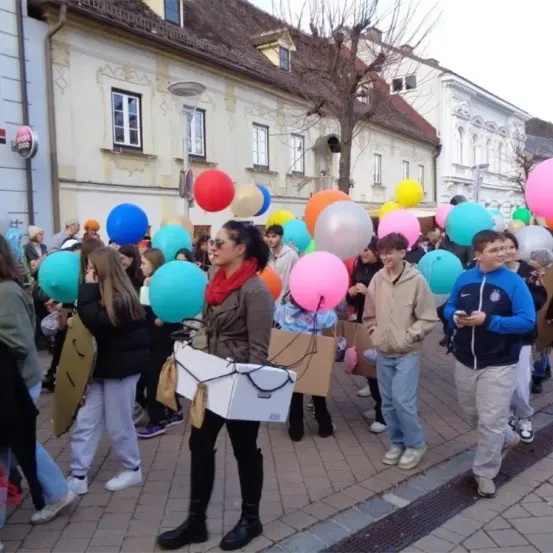 Eine Gruppe von Menschen bei einer Straßenparade mit bunten Luftballons, in verschiedenen Kleidungsstücken und mit Kisten, vor Gebäuden mit Fenstern und einem Baum.