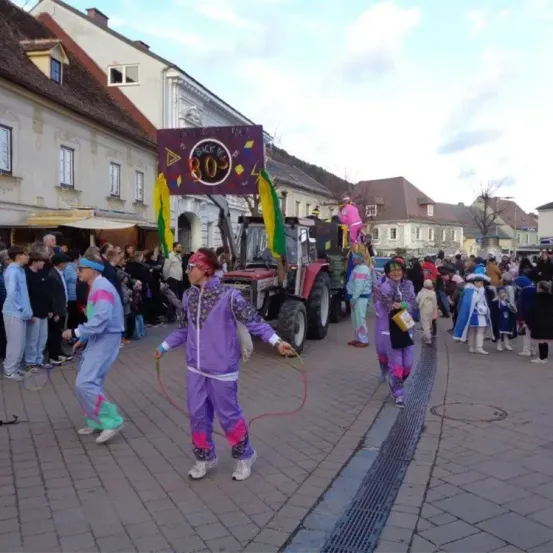 Ein Umzug mit Menschen in bunten Kostümen und einem Traktor in der Mitte. Menschen beobachten von der Straßenseite.