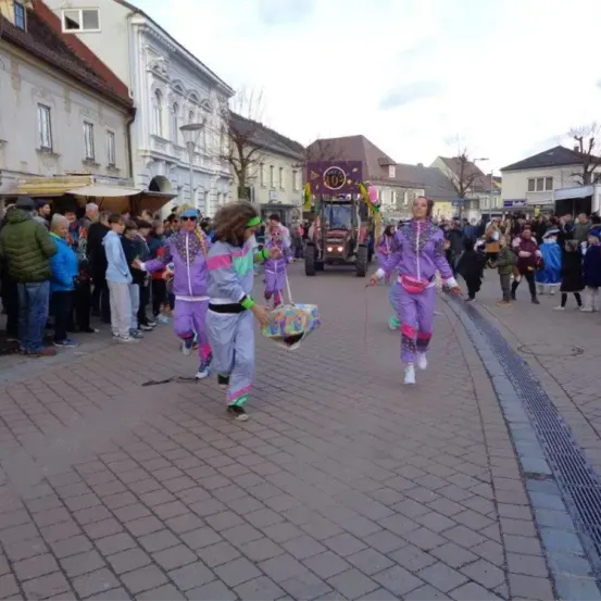 Eine Gruppe von Menschen in bunten Kostümen läuft auf einer Straße, mit einem Traktor und einer zuschauenden Menge. Gebäude säumen die Straße.