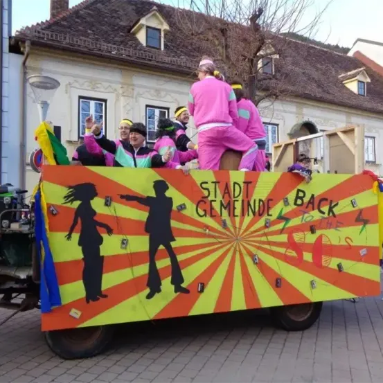 Ein bunter Umzugswagen mit Menschen in rosa und grünen Kostümen, die winken. Der Wagen hat einen kräftigen gelben und orangefarbenen Hintergrund mit Silhouetten und dem Text 'Stadt Gemeinde Back To The 80s.'