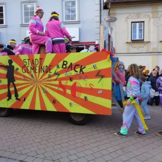 Ein Float mit Menschen in rosa Anzügen fährt durch die Straße. Der Float sagt Stadt Gemeinde Zurück in die Zukunft.