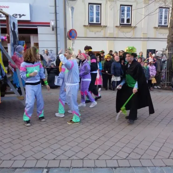 Eine Gruppe von Menschen in farbenfrohen Kostümen und Hüten führt auf der Straße eine Aufführung durch. Sie halten Requisiten. Im Hintergrund befindet sich ein Gebäude mit vielen Fenstern und einem Baum.