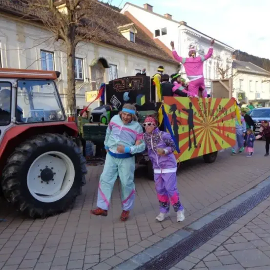 Ein Mann und eine Frau in Retro-Kleidung der 80er stehen vor einem Traktor, während ein Wagen mit Leuten in Pink dahinter ist.